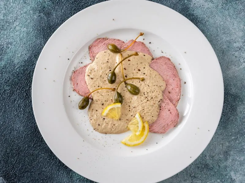 Thin pale-pink veal slices under a creamy tuna sauce with caper berries, lemon wedges and black pepper on a white plate