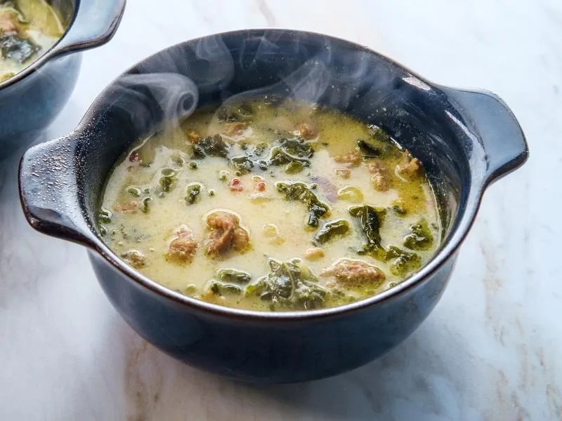 Creamy pale soup with pieces of sausage, sliced potatoes and leafy kale in a dark handled bowl