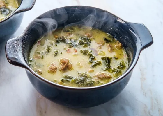 Creamy pale soup with pieces of sausage, sliced potatoes and leafy kale in a dark handled bowl